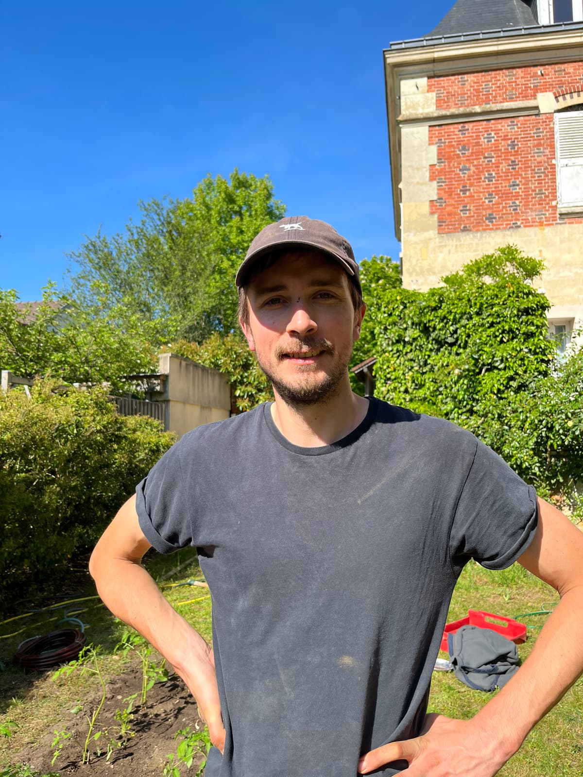 Portrait de Léo debout au potager, t-shirt gris, jardin et maison en arrière-plan.